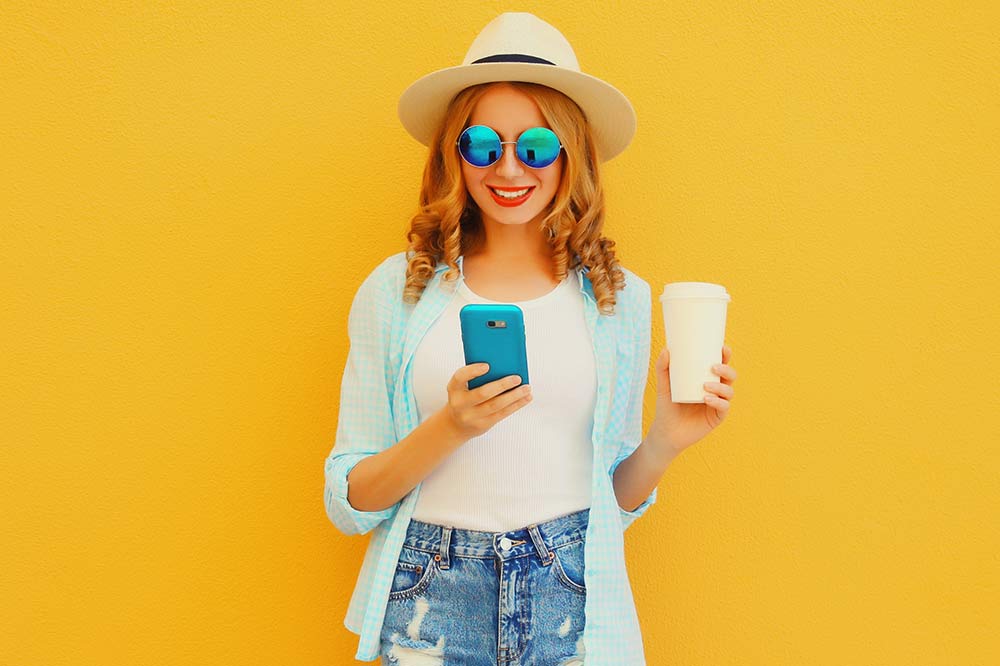 Happy person with sunglasses and hat holding a phone and coffee with yellow background